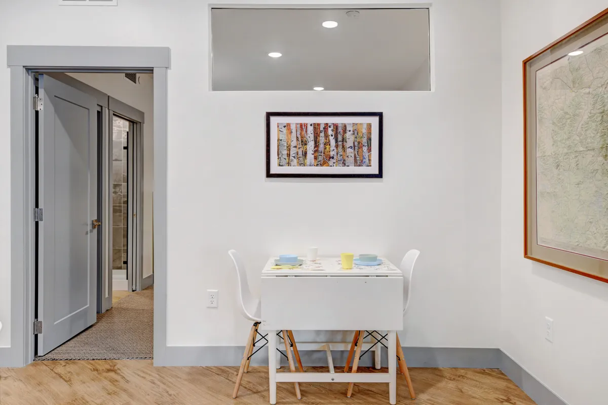 A small, intimate dining table set with colorful tableware, framed artwork, and a nearby doorway that suggests a connection to the outside or other living spaces. The overall vibe is cozy and suited for private meals or quiet conversation.