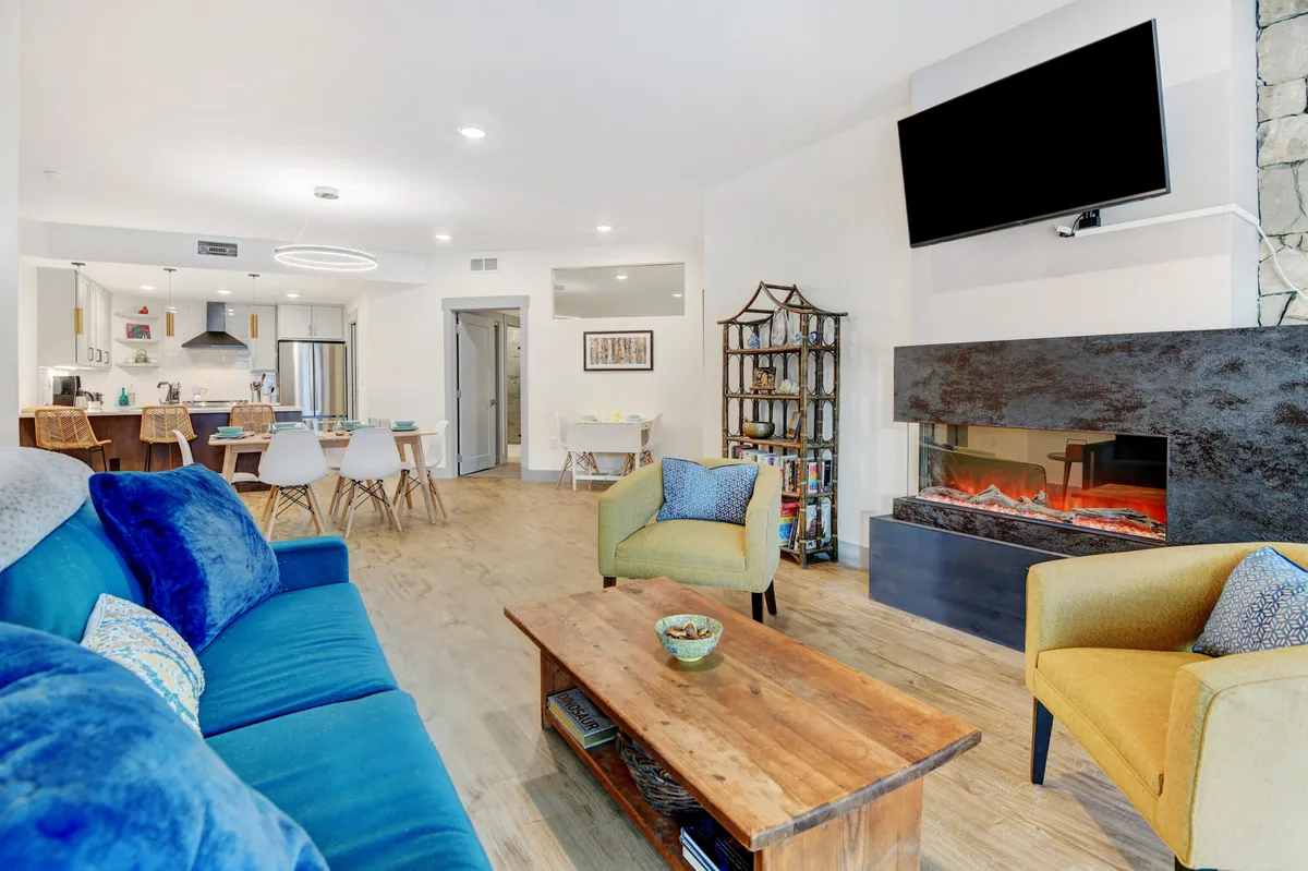 This broad interior shot displays the living room, with a blue couch, wooden coffee table, a fireplace, and the dining area in the background. The scene is cozy, with the fireplace ready for après-ski relaxation or game nights. The open layout visually ties together the living, dining, and kitchen spaces.