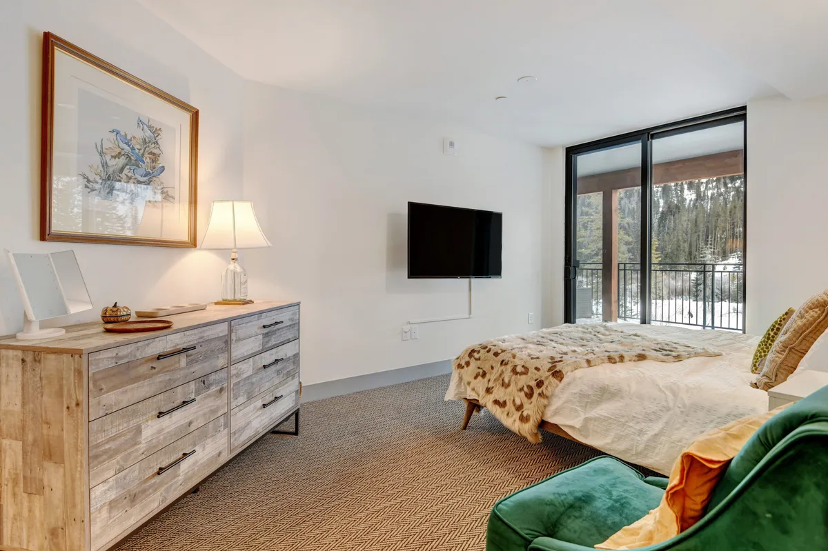 A cozy, well-lit bedroom featuring a neatly made bed with a soft throw blanket, a green armchair perfect for reading or relaxing, a wooden dresser with a lamp, and a mounted television. The space appears serene, organized, and immaculately maintained, with no traces of animal presence.