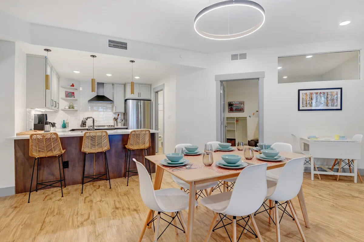 This image showcases the transition from kitchen to dining area, with a dining table elegantly set with plates and cutlery and a kitchen island visible in the background. A modern light fixture and a welcoming, contemporary mood set the tone. The space looks ready for shared meals or celebratory occasions.