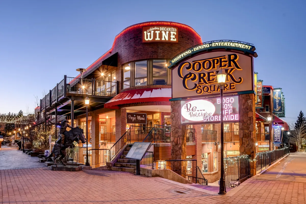 This exterior shot captures the lively Cooper Creek Square at dusk, with illuminated storefronts, decorative lights, and people strolling by Peggy's Wine entrance. The festive energy and abundance of dining and shopping options are visible, showcasing the downtown's vibrant atmosphere.
