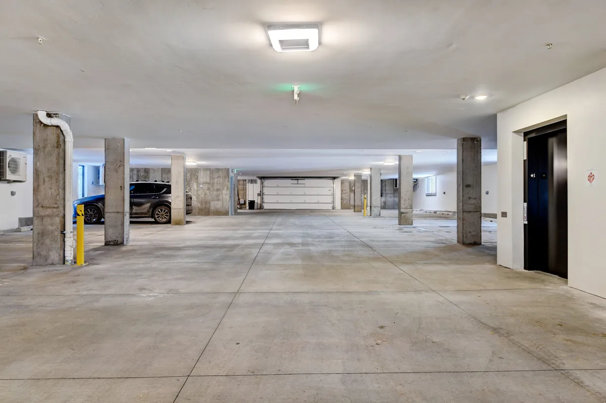 A practical, well-lit underground parking garage featuring empty spaces, concrete pillars, an elevator entrance, and a parked vehicle. The scene conveys ease of access, security, and convenience for residents.