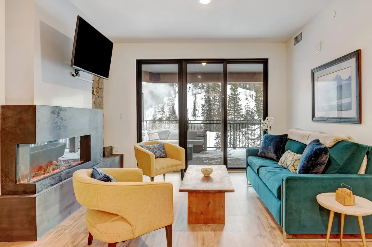 This inviting living room features a cozy fireplace, plush sofa, and expansive floor-to-ceiling windows framing breathtaking Winter Park mountain views. Natural light floods the space, creating a warm ambiance perfect for relaxing, sipping coffee, or gathering with friends and family while snow falls gently outside.