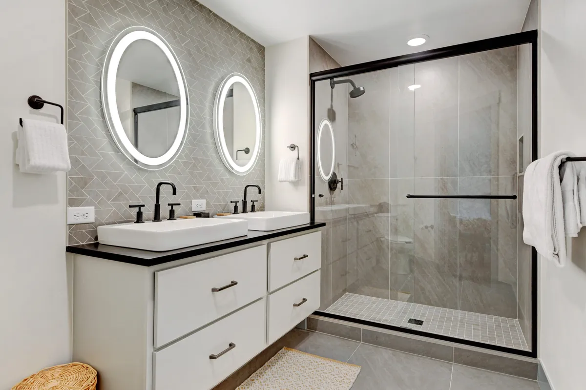 A spa-inspired bathroom with double sinks featuring modern fixtures, round LED-lit mirrors, and a glass-enclosed shower. The clean lines and upscale finishes evoke a sense of relaxation and luxury.