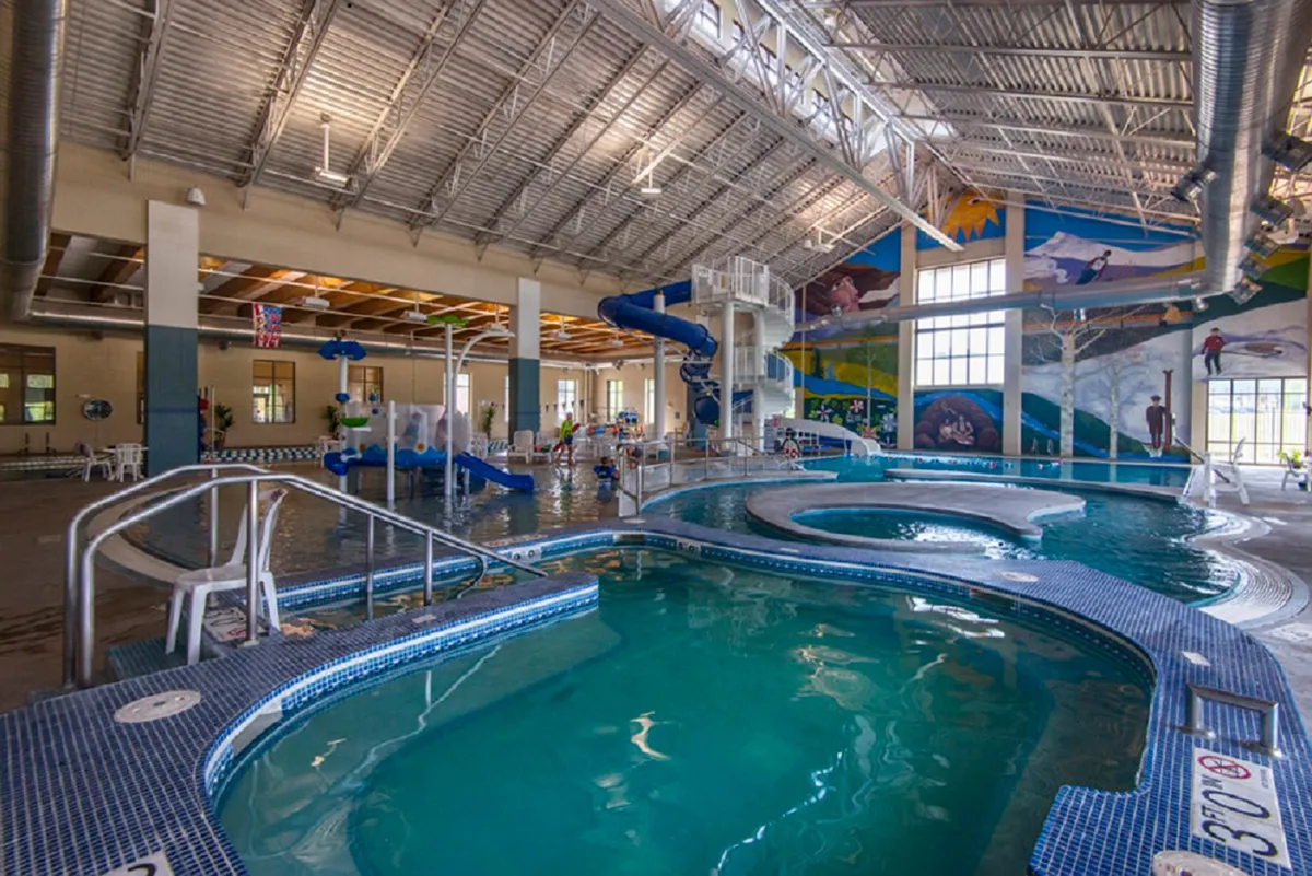 The scene captures the vibrant Grand Park Rec Center interior, featuring water slides, a large swimming pool, colorful murals, and children joyfully playing. The energy and family-friendly atmosphere are palpable, reflecting the resort-style amenities accessible to guests.