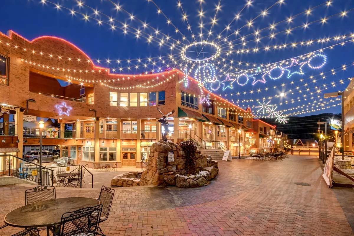 The scene showcases a lively exterior plaza illuminated by decorative lights, featuring a central fountain and surrounded by shops. The vibrant atmosphere hints at bustling nightlife, outdoor dining, and the convenience of walkable local attractions in the downtown area.