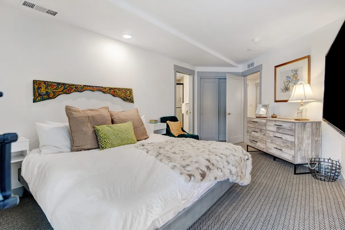 A cozy bedroom featuring a well-made bed with a decorative throw, a dresser with a softly glowing lamp, and a piece of art on the wall. The space feels inviting and serene, ideal for relaxation and restful sleep.