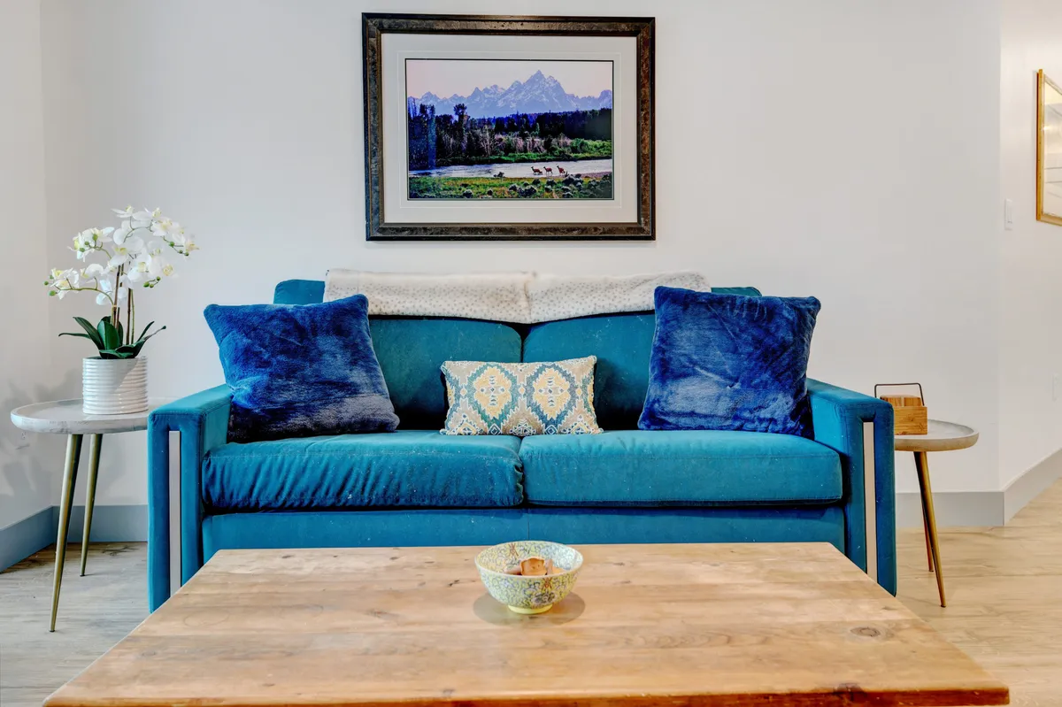 This image shows a bright, inviting living room with a stylish blue couch accented by colorful pillows. Above the couch hangs a vibrant piece of artwork, and a clean wooden coffee table sits in the foreground. The scene exudes comfort, calm, and a sense of freshness, with no signs of pet presence, fur, or clutter.